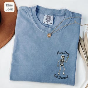 May include: A blue jean colored t-shirt with a crew neck and short sleeves. The shirt features a skeleton graphic holding a cocktail and the text "Bone Dry but Buzzed". A gold necklace with a pendant is draped on the shirt.