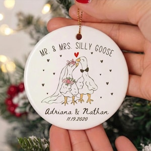 May include: A white ceramic Christmas ornament with a gold string. The ornament features a cartoon illustration of two geese, one wearing a veil and holding flowers, the other wearing a bow tie. Text reads "MR. & MRS. SILLY GOOSE" and the names "Adriana & Nathan" with the date "11.19.2020".