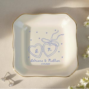 May include: A white ceramic trinket dish with a gold rim. The dish features a blue heart-shaped design with the letters "a" and "n", along with the names "Adriana & Nathan" and the date "11.19.2020".