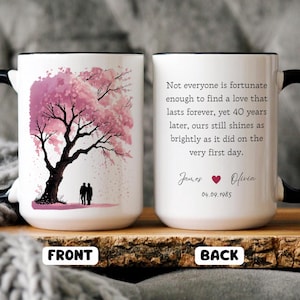 May include: White ceramic mug with a black handle and rim. The front displays a pink cherry blossom tree and two silhouettes. The back has a quote about love, names, a heart, and a date.