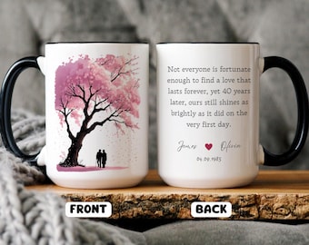 May include: White ceramic mug with a black handle and rim. The front features a pink cherry blossom tree with two silhouettes. The back has a quote about love, names, a heart, and a date.