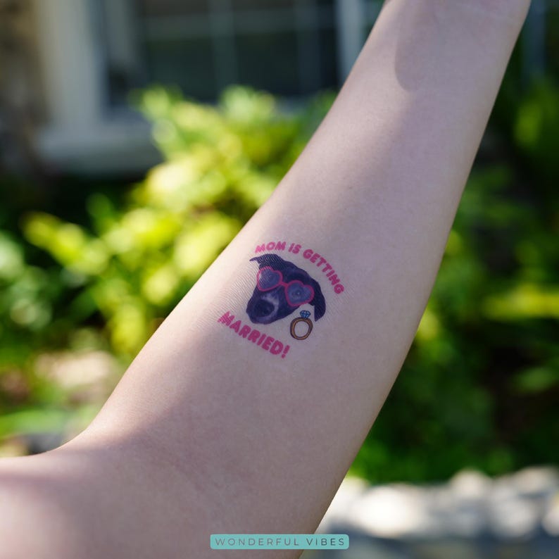 Peut inclure: Un tatouage temporaire repr&eacute;sentant un chien noir portant des lunettes en forme de c&oelig;ur et une alliance. Le texte rose "MOM IS GETTING MARRIED!" est visible. Le tatouage est sur le bras d'une personne.
