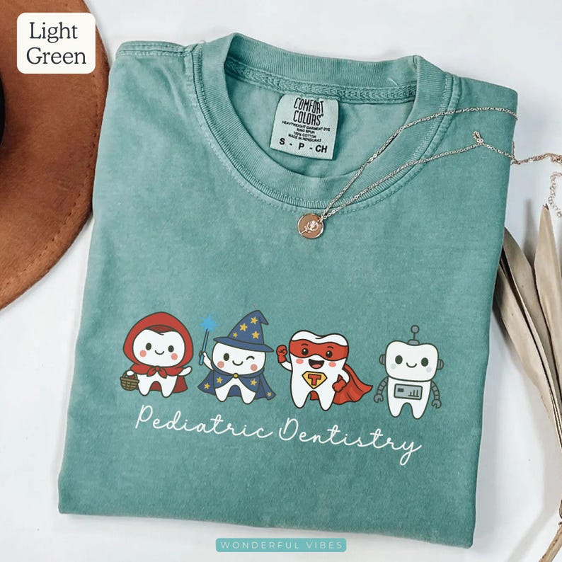 K&ouml;nnte beinhalten: Hellgr&uuml;nes T-Shirt mit dem Text "Pediatric Dentistry" und Cartoonfiguren, die als Rotk&auml;ppchen, Zauberer, Superheld und Roboter verkleidet sind. Eine silberne Halskette mit Anh&auml;nger liegt auf dem Shirt.