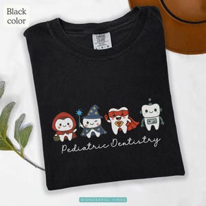 K&ouml;nnte beinhalten: Schwarzes T-Shirt mit vier Cartoon-Z&auml;hnen, die als Rotk&auml;ppchen, Zauberer, Superheld und Roboter verkleidet sind. Darunter steht in Schreibschrift "Pediatric Dentistry".
