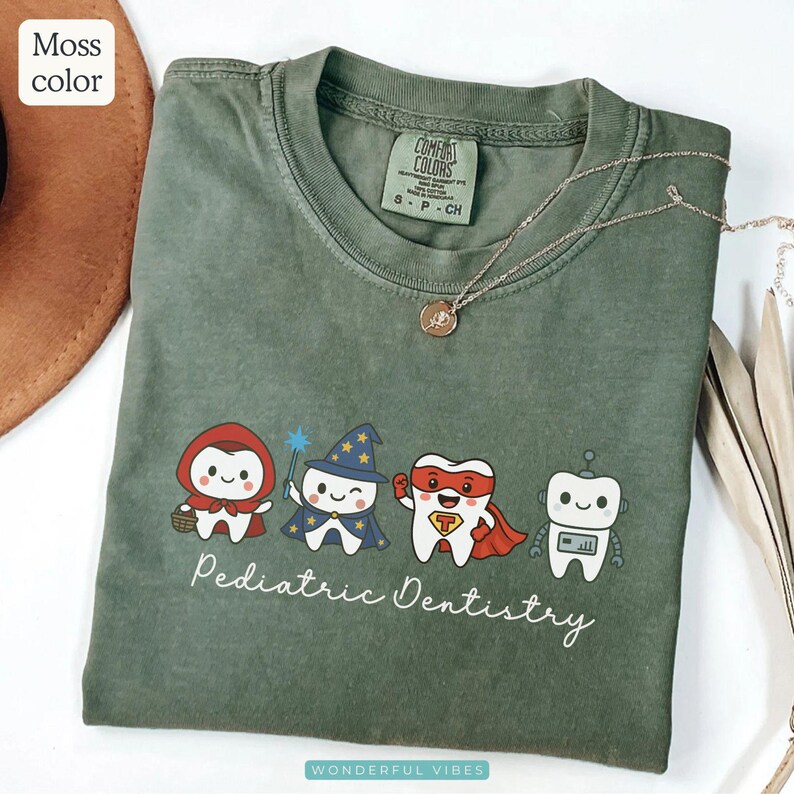 K&ouml;nnte beinhalten: Ein moosgr&uuml;nes T-Shirt mit einer Grafik von Zeichentrickz&auml;hnen, die als Charaktere verkleidet sind, darunter Rotk&auml;ppchen, ein Zauberer, ein Superheld und ein Roboter. Die Worte "Pediatric Dentistry" stehen darunter. Eine goldene Halskette liegt &uuml;ber dem Shirt.