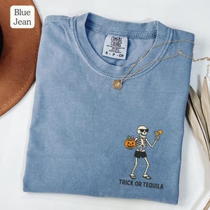 May include: Blue jean Comfort Colors t-shirt with a skeleton graphic holding a pumpkin and a cocktail. The text "Trick or Tequila" is printed below the graphic. A gold necklace is draped across the shirt.