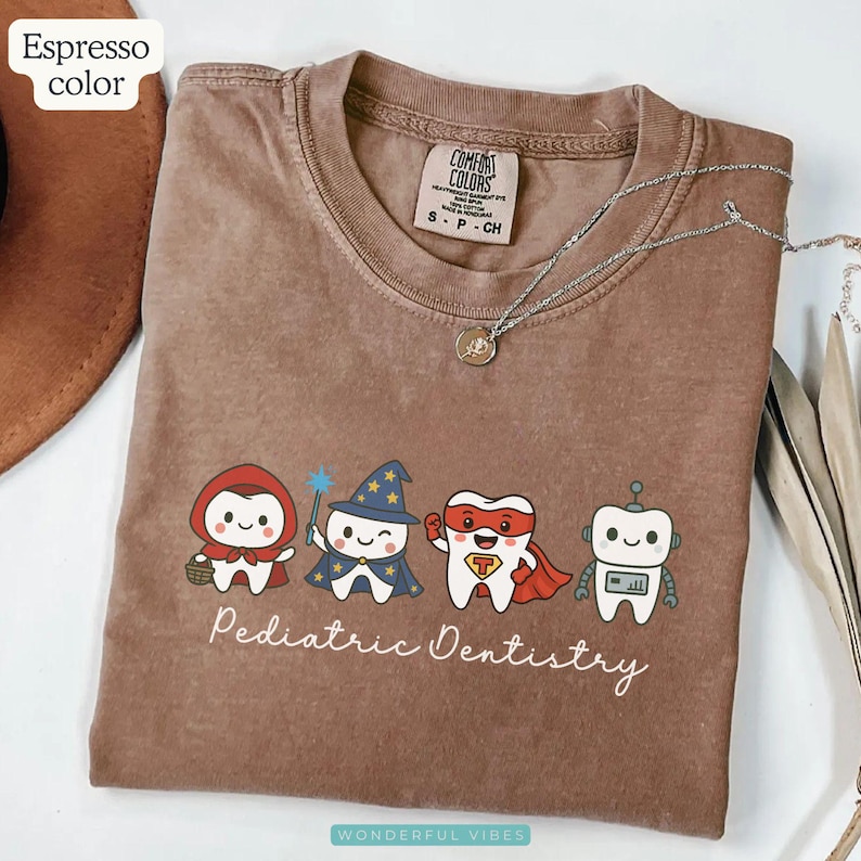 K&ouml;nnte beinhalten: Espressofarbenes T-Shirt mit einer Grafik von Zeichentrickz&auml;hnen, die als Charaktere verkleidet sind. Die Charaktere sind Rotk&auml;ppchen, ein Zauberer, ein Superheld und ein Roboter. Darunter steht der Text "Pediatric Dentistry".