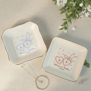 May include: Two off-white ceramic ring dishes with gold trim. Each dish features a heart design with initials and a date. One dish has blue accents, the other red. A gold necklace and earrings are also visible.