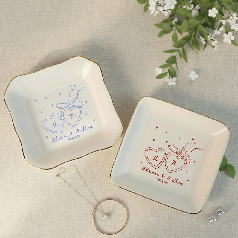 May include: Two off-white ceramic ring dishes with gold trim. Each dish features a heart design with initials and a date. One dish has blue accents, the other red. A gold necklace and earrings are also visible.