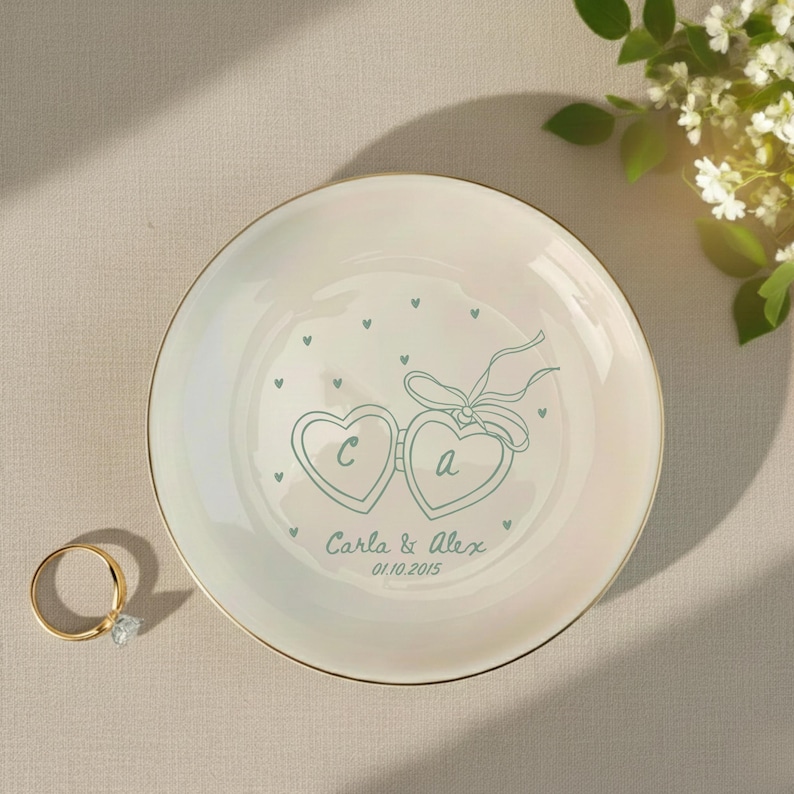 May include: A white ceramic ring dish with a gold rim. The dish features a teal heart-shaped design with the letters "C" and "A", a bow, and the names "Carla & Alex" with the date "01.10.2015". A gold ring with a diamond is on the left.
