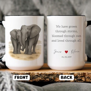 Puede incluir: Taza de café de cerámica blanca con asa y borde negros. La parte delantera de la taza presenta una ilustración de dibujos animados de dos elefantes. La parte posterior de la taza tiene el texto "We have grown through storms, bloomed through sun and loved through all." con los nombres "James" y "Olivia" y la fecha "04.09.2011".