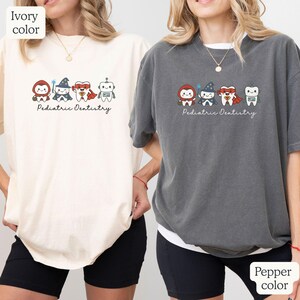 K&ouml;nnte beinhalten: Elfenbein- und pfefferfarbene T-Shirts mit einer Cartoon-Grafik von Superhelden-Z&auml;hnen. Die Grafik zeigt einen Zahn als Superheld, einen Zauberer und einen Roboter. Der Text "Pediatric Dentistry" steht darunter.