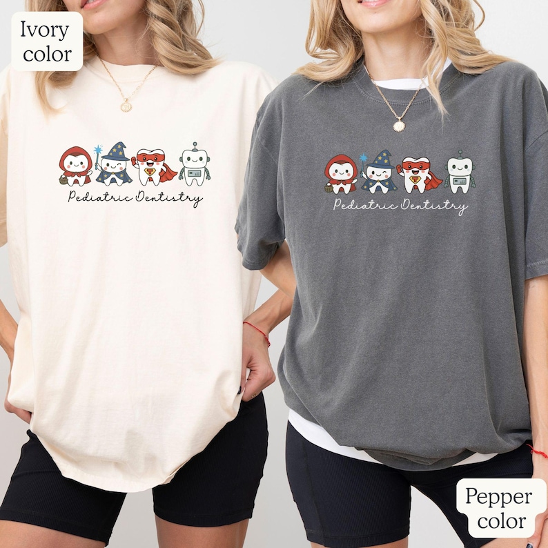 K&ouml;nnte beinhalten: Elfenbein- und pfefferfarbene T-Shirts mit einer Cartoon-Grafik von Superhelden-Z&auml;hnen. Die Grafik zeigt einen Zahn als Superheld, einen Zauberer und einen Roboter. Der Text "Pediatric Dentistry" steht darunter.