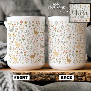 May include: White ceramic mug with a black handle and rim, featuring a floral and duck pattern. The design includes various flowers and ducks in shades of yellow, green, and brown. The back of the mug has the name "Olivia" in a floral design.
