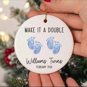 May include: A white ceramic Christmas ornament with the text "MAKE IT A DOUBLE" and two pairs of blue baby footprints. The ornament also includes the text "Williams Twins" and the date "FEBRUARY 2026".