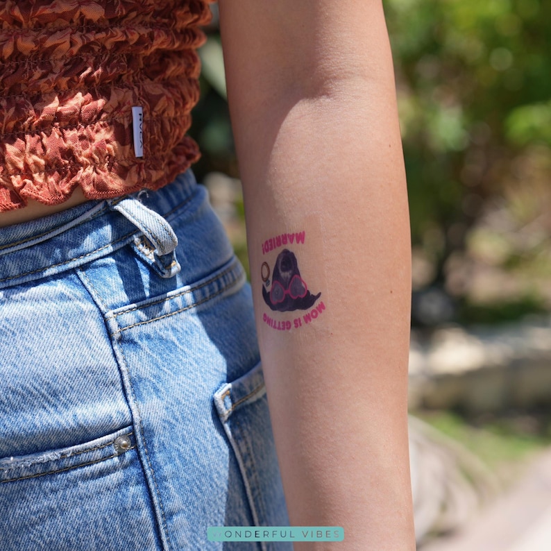 Peut inclure: Un tatouage temporaire sur un bras avec le texte "MOM IS GETTING MARRIED!" en rose. Le tatouage repr&eacute;sente une image de dessin anim&eacute; d'une mari&eacute;e avec des lunettes roses et un voile. La personne porte un haut marron et un jean bleu.