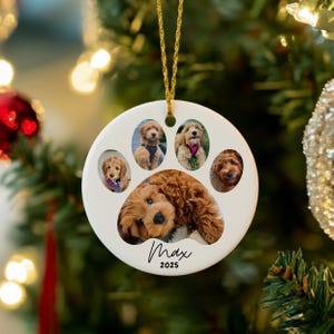 May include: A white ceramic Christmas ornament shaped like a paw print, featuring five photos of a dog. The dog is brown with curly fur. The name "Max" and the year "2025" are written in black script. A gold string is attached for hanging.