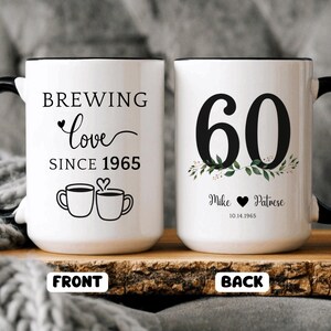 Peut inclure: Deux mugs en céramique blancs avec des anses et des bords noirs. L'un des mugs indique "Brewing Love Since 1965" avec des illustrations de tasses à café. L'autre mug porte un grand "60" avec des accents floraux et des noms et une date.