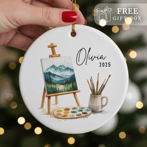 May include: White ceramic ornament with a gold string, featuring a watercolor painting of a mountain landscape on a small easel. Includes a palette of paints, paintbrushes in a mug, and the name "Olivia 2025".