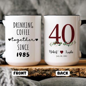 May include: White ceramic mug with black handle and rim. The front of the mug reads "DRINKING COFFEE together SINCE 1985". The back of the mug has a large burgundy "40" with floral accents and the names "Robert & Linda" and the date "July 28, 1985".