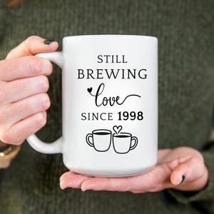 Pu&ograve; includere: Tazza in ceramica bianca con la scritta "STILL BREWING Love SINCE 1998" in corsivo nero. La tazza presenta due illustrazioni di tazze da caff&egrave; con un cuore sopra. La tazza &egrave; tenuta da una persona.