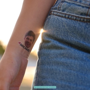 Peut inclure: Un tatouage temporaire repr&eacute;sentant le visage souriant d'un homme avec le nom "MICHAEL" et un c&oelig;ur. Le tatouage est sur un poignet, avec le soleil en arri&egrave;re-plan. La personne porte un jean bleu.