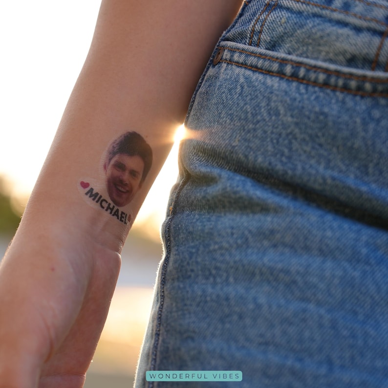 Peut inclure: Un tatouage temporaire repr&eacute;sentant le visage souriant d'un homme avec le nom "MICHAEL" et un c&oelig;ur. Le tatouage est sur un poignet, avec le soleil en arri&egrave;re-plan. La personne porte un jean bleu.