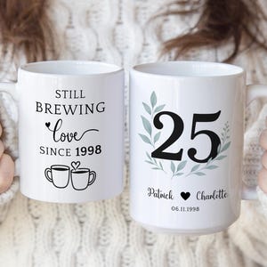 Pu&ograve; includere: Due tazze in ceramica bianca. Una tazza recita "Still Brewing Love Since 1998" con illustrazioni di tazze da caff&egrave;. L'altra tazza presenta il numero 25 con nomi e una data, per celebrare un anniversario.