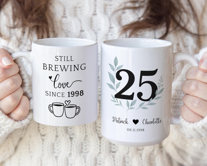 Pu&ograve; includere: Due tazze in ceramica bianca. Una tazza recita "Still Brewing Love Since 1998" con illustrazioni di tazze da caff&egrave;. L'altra tazza presenta il numero 25 con nomi e una data, per celebrare un anniversario.