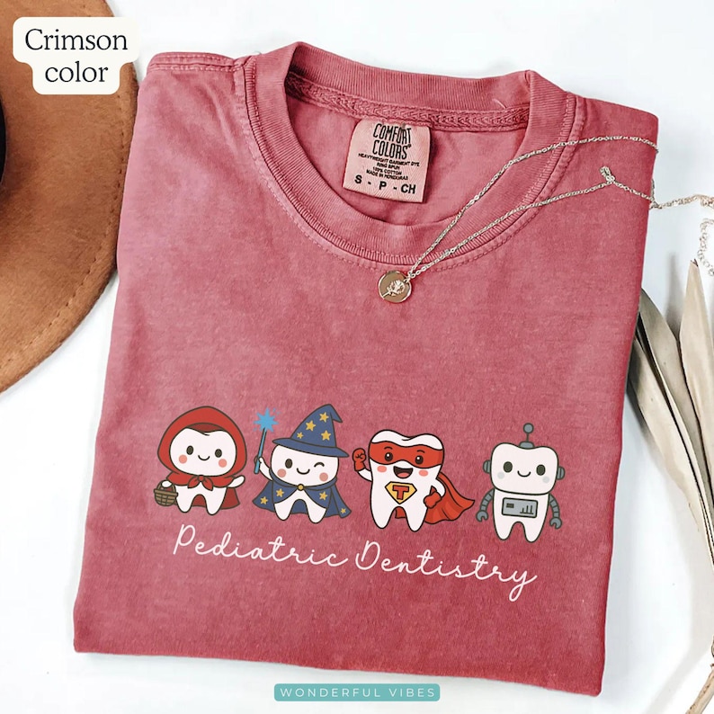 K&ouml;nnte beinhalten: Karmesinrotes T-Shirt mit Rundhalsausschnitt und dem Text "Pediatric Dentistry". Das Shirt zeigt Zeichentrickz&auml;hne, die als Charaktere verkleidet sind. Eine silberne Halskette mit Anh&auml;nger ist auf dem Shirt drapiert.
