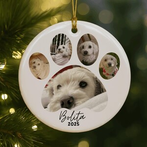 May include: White ceramic paw print ornament featuring five photos of a white dog. The ornament has a gold string for hanging and the name "Bolita" and the year "2025" are written in black script.