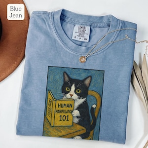 May include: Blue jean colored t-shirt with a graphic of a black and white cat reading a book titled "Human Manipulation 101." The shirt has a crew neck and short sleeves. A gold necklace with a pendant is draped across the shirt.