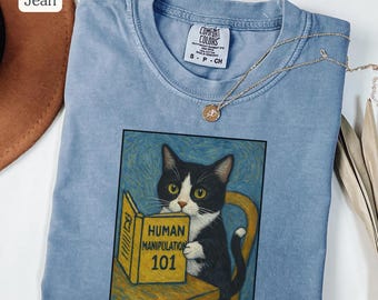 May include: Blue jean colored t-shirt with a graphic of a black and white cat reading a book titled "Human Manipulation 101." The shirt has a crew neck and short sleeves. A gold necklace with a pendant is draped across the shirt.