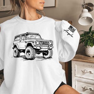 May include: White sweatshirt with a black and white illustration of a vintage Bronco on the front. The sweatshirt has a black American flag patch on the sleeve with the word "Angela" written on it.