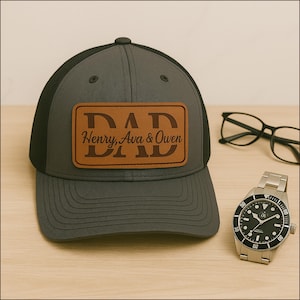 Może przedstawiać: Szara i czarna czapka typu trucker z brązową skórzaną naszywką z napisem "DAD" nad imionami "Henry, Ava & Owen". Widoczny jest również srebrny zegarek i czarne okulary.