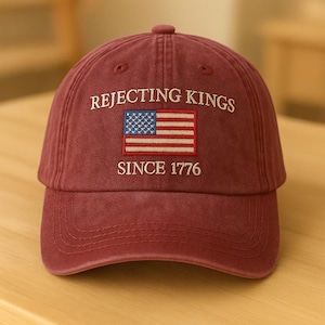 Może przedstawiać: Bordowa czapka bejsbolowa z napisem "REJECTING KINGS" nad haftowaną amerykańską flagą i napisem "SINCE 1776". Czapka ma zakrzywiony daszek i regulowany pasek.