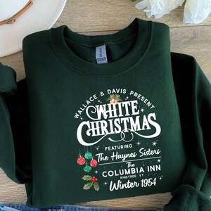 May include: A dark green sweatshirt featuring the words "WHITE CHRISTMAS" in white script. The design includes the text "WALLACE & DAVIS PRESENT" and "FEATURING The Haynes Sisters The COLUMBIA INN PINETREE, VT Winter 1954". The design includes festive ornaments and holly.