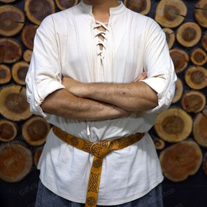 May include: A white linen shirt with a lace-up neckline and long sleeves. The shirt is worn with a brown leather belt with a Celtic knot design.