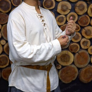 May include: A white linen shirt with a lace-up front and long sleeves. The shirt has a button on the left cuff and a brown leather belt with a Celtic knot design.