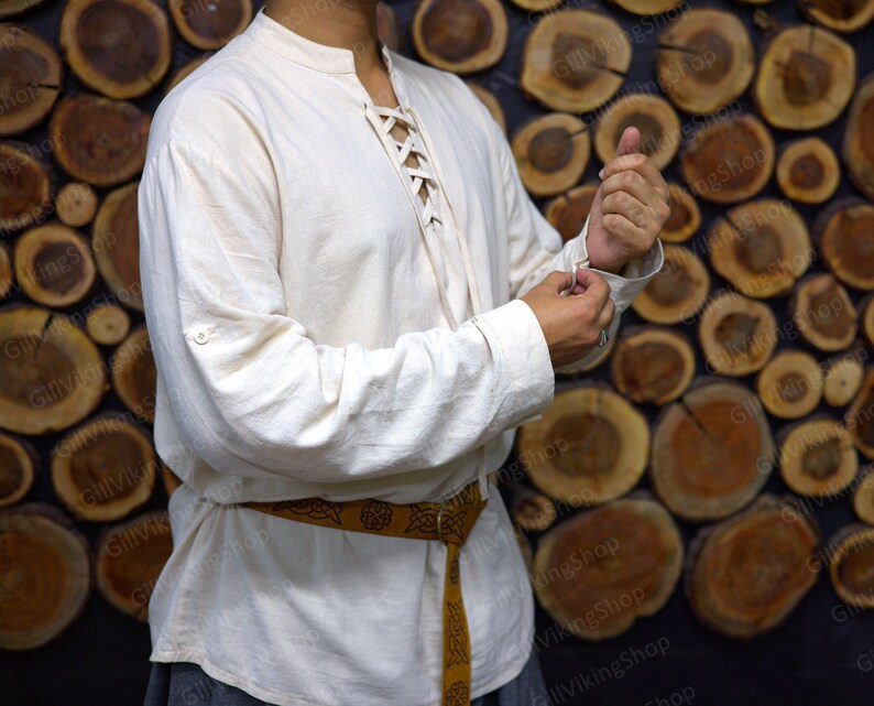 May include: A white linen shirt with a lace-up front and long sleeves. The shirt has a button on the left cuff and a brown leather belt with a Celtic knot design.