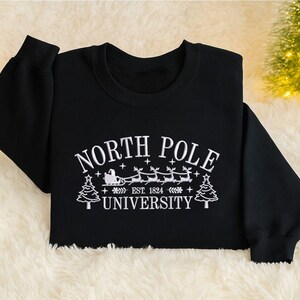 以下が含まれることがあります： 白い文字で「NORTH POLE UNIVERSITY EST. 1824」と書かれた黒いクルーネックのスウェットシャツ。サンタのそりとトナカイのグラフィックが描かれています。スウェットシャツはふわふわの白い表面にあり、背景には小さなクリスマスツリーがあります。