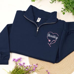 May include: Navy blue quarter-zip sweatshirt with a pink heart-shaped stethoscope design and the name "Bentley". The sweatshirt has a ribbed collar and cuffs. The design is embroidered on the left chest.