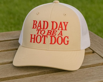 May include: A tan baseball cap with a white mesh back. The front of the cap features the red embroidered text, "BAD DAY TO BE A HOT DOG". The cap is sitting on a wooden surface.