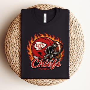 Puede incluir: Camiseta negra con un diseño de casco y balón de fútbol americano rojo, con el texto "LET'S GO" y "Chiefs". El diseño incluye llamas alrededor del casco y el balón. La camiseta está doblada sobre una superficie tejida.