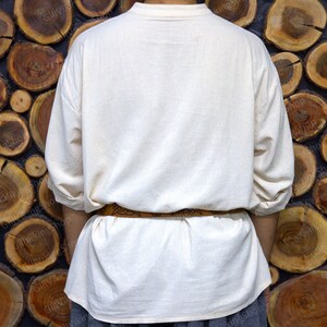 May include: A white linen tunic with long sleeves and a brown leather belt. The tunic is worn by a person standing in front of a wall made of stacked wooden rounds.