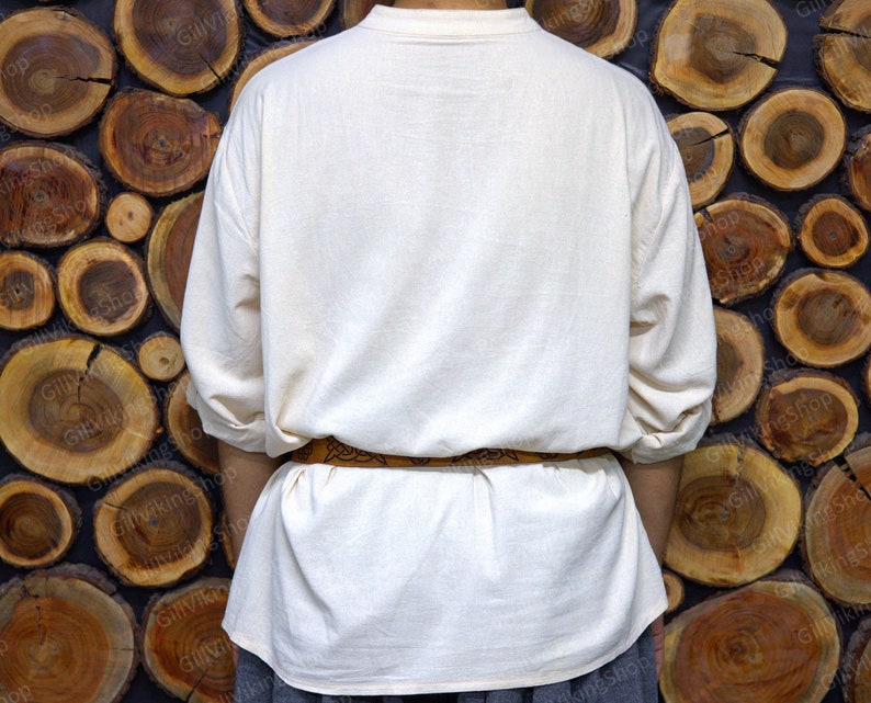 May include: A white linen tunic with long sleeves and a brown leather belt. The tunic is worn by a person standing in front of a wall made of stacked wooden rounds.