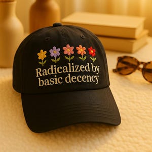 Op de afbeelding: Zwarte baseballcap met geborduurde bloemen in geel, paars, roze, oranje en rood. De cap heeft de tekst "Radicalized by basic decency" in witte letters. Een stijlvol accessoire voor dagelijks gebruik.