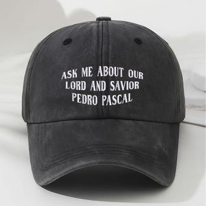 May include: A dark gray baseball cap with the embroidered text "ASK ME ABOUT OUR LORD AND SAVIOR PEDRO PASCAL" in white. The cap has a curved brim and a button on top. The hat is made of a soft, worn material.