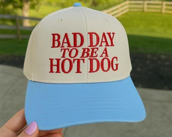May include: A baseball cap with a cream-colored crown and a light blue brim. The cap features the embroidered phrase "BAD DAY TO BE A HOT DOG" in red lettering. The cap has a rounded top and a curved brim.