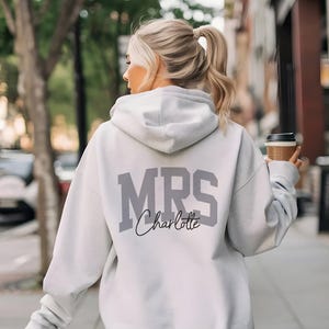 May include: A light gray hoodie with the word "MRS" in large, gray block letters and the name "Charlotte" in a script font. The person is walking down a city street and holding a coffee cup.